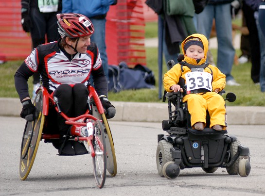 Image of Chris in a racing chair with a child in a motorized wheelchair