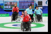 Jim Armstrong curling with Ina Forrest holding his chair and Sonia Gaudet in the background on the ice
