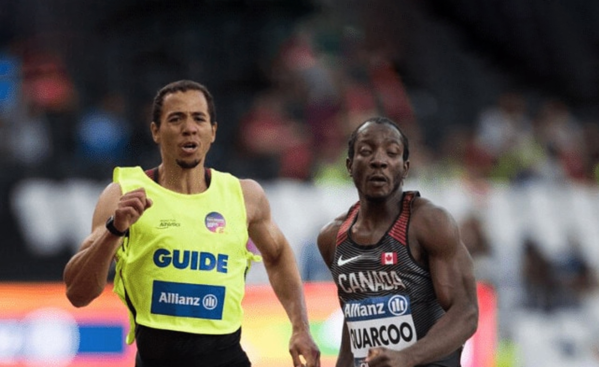 L’athlète paralympique canadien George Quarcoo sprinte aux côtés de son guide voyant lors d’une course. Quarcoo porte un uniforme noir de l’équipe canadienne avec des accents rouges, affichant son nom et un drapeau canadien sur sa poitrine. Son guide, vêtu d’un gilet jaune vif marqué « GUIDE », court à ses côtés. Les deux athlètes affichent une concentration intense alors qu’ils concourent dans un stade avec une foule floue en arrière-plan.