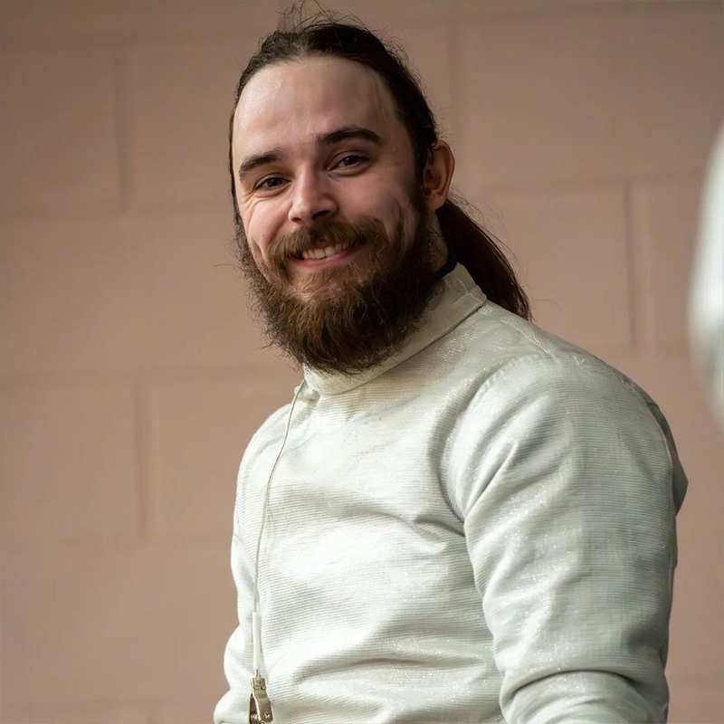 Photo en gros plan de Ryan Rousell, escrimeur canadien en fauteuil roulant, souriant à la caméra en portant sa tenue d'escrime.