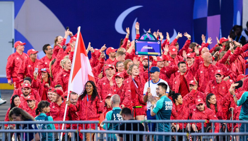 L'équipe paralympique canadienne, vêtue d'uniformes rouges, entre dans le stade lors de la cérémonie d'ouverture des Jeux paralympiques de Paris 2024. Les membres de l'équipe saluent et acclament, avec le drapeau canadien et le panneau de l'équipe tenus fièrement à l'avant.