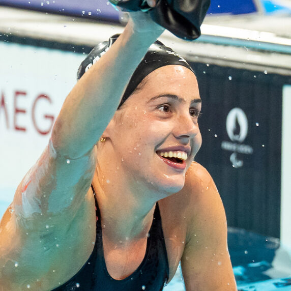 La nageuse paranatation canadienne Aurélie Rivard sourit de joie en levant le bras pour célébrer après avoir terminé une course aux Jeux paralympiques. Elle est encore dans la piscine, de l'eau coule de son corps, et elle porte un bonnet de bain noir et un maillot de bain noir. En arrière-plan, on voit le système de chronométrage avec "Omega" visible.