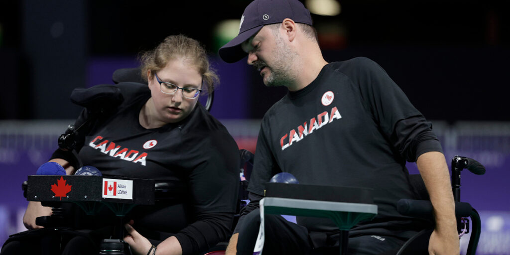 L'athlète canadienne de boccia Alison Levine, portant un maillot noir de l'équipe du Canada, est assise dans son fauteuil roulant et est concentrée sur sa rampe de boccia. Un entraîneur ou assistant, également vêtu d'un maillot noir du Canada, se penche pour l'aider ou lui donner des conseils pendant le match. Plusieurs balles de boccia sont visibles sur la rampe, et les deux personnes sont concentrées sur le jeu.