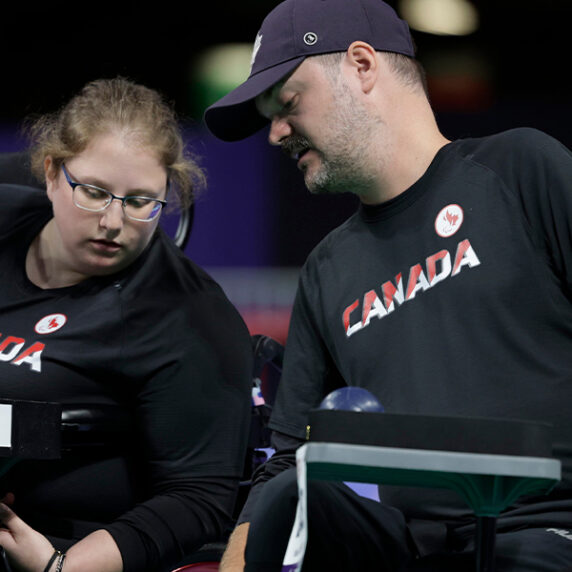 L'athlète canadienne de boccia Alison Levine, portant un maillot noir de l'équipe du Canada, est assise dans son fauteuil roulant et est concentrée sur sa rampe de boccia. Un entraîneur ou assistant, également vêtu d'un maillot noir du Canada, se penche pour l'aider ou lui donner des conseils pendant le match. Plusieurs balles de boccia sont visibles sur la rampe, et les deux personnes sont concentrées sur le jeu.