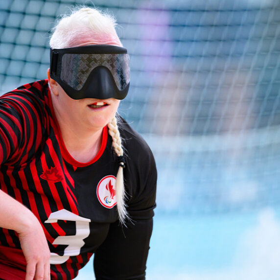 Whitney Bogart, joueuse canadienne de goalball, en action lors d'un match. Elle porte un maillot de l'équipe noir et rouge avec le numéro 3, ainsi qu'un masque oculaire noir, un équipement standard en goalball. Whitney est accroupie sur le terrain, concentrée et prête à défendre.