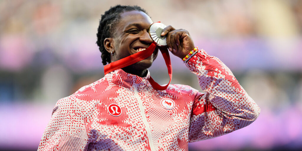 L'image montre Jesse Zesseu, un para-athlète canadien, souriant avec fierté en mordant sa médaille d'argent lors de la cérémonie de remise des médailles aux Jeux paralympiques de Paris 2024. Il porte une veste rouge et blanche à motifs avec les logos de l'équipe paralympique canadienne et de Lululemon visibles. Zesseu tient la médaille d'une main, avec le ruban rouge pendu. L'arrière-plan est flou, mettant en valeur son expression joyeuse.