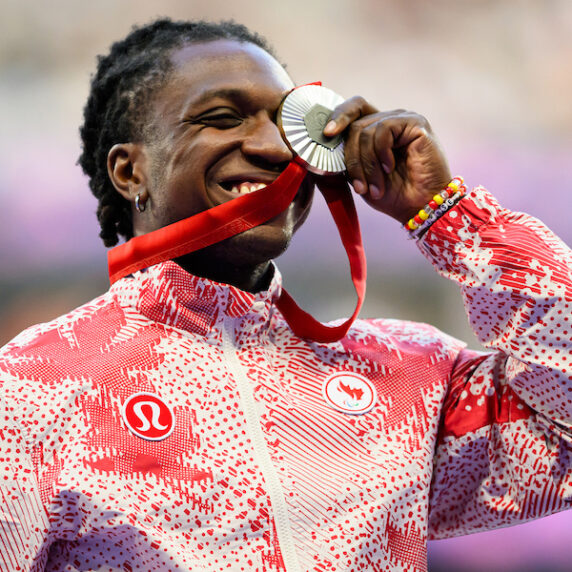 L'image montre Jesse Zesseu, un para-athlète canadien, souriant avec fierté en mordant sa médaille d'argent lors de la cérémonie de remise des médailles aux Jeux paralympiques de Paris 2024. Il porte une veste rouge et blanche à motifs avec les logos de l'équipe paralympique canadienne et de Lululemon visibles. Zesseu tient la médaille d'une main, avec le ruban rouge pendu. L'arrière-plan est flou, mettant en valeur son expression joyeuse.