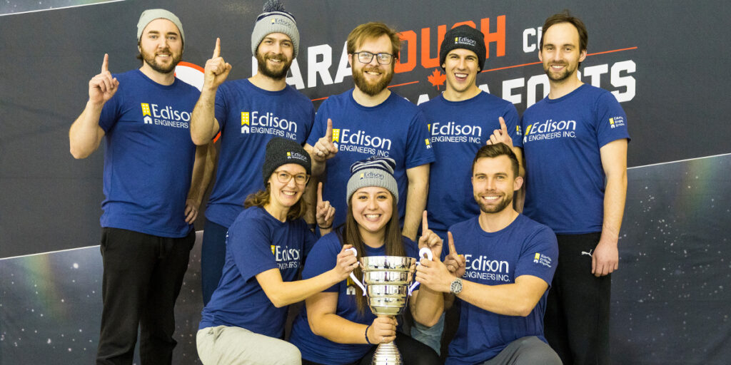 Un groupe de huit personnes portant des t-shirts bleus assortis de Edison Engineers et des bonnets d'hiver posent ensemble avec un grand trophée. Ils sourient et lèvent un doigt pour indiquer qu'ils sont numéro un, célébrant leur victoire lors de l'événement ParaTough Cup.