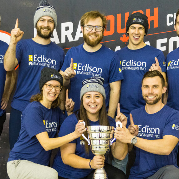 Un groupe de huit personnes portant des t-shirts bleus assortis de Edison Engineers et des bonnets d'hiver posent ensemble avec un grand trophée. Ils sourient et lèvent un doigt pour indiquer qu'ils sont numéro un, célébrant leur victoire lors de l'événement ParaTough Cup.