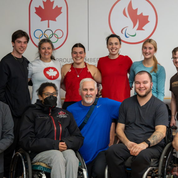 L’image montre un groupe de personnes diverses, certaines debout et d’autres en fauteuil roulant, posant ensemble devant un mur affichant les logos des équipes olympique et paralympique canadiennes. Tous sourient, créant une ambiance positive et inclusive. Le cadre semble être une installation d’entraînement ou une activité de cohésion d’équipe, reflétant un fort esprit de camaraderie et de travail d’équipe.