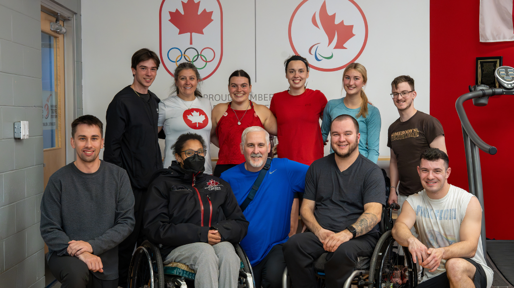 L’image montre un groupe de personnes diverses, certaines debout et d’autres en fauteuil roulant, posant ensemble devant un mur affichant les logos des équipes olympique et paralympique canadiennes. Tous sourient, créant une ambiance positive et inclusive. Le cadre semble être une installation d’entraînement ou une activité de cohésion d’équipe, reflétant un fort esprit de camaraderie et de travail d’équipe.