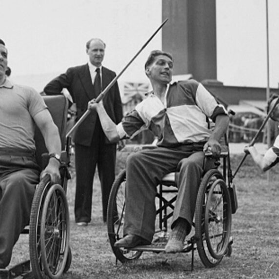 Photo en noir et blanc d'athlètes en fauteuil roulant participant à une épreuve de javelot. Trois hommes en fauteuil roulant se préparent à lancer des javelots dans un cadre extérieur, observés par des spectateurs en arrière-plan.