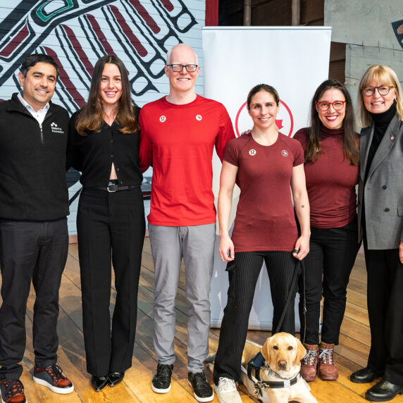 Un groupe de huit personnes, comprenant des athlètes paralympiques, des représentants de fondations et un chien-guide, est debout côte à côte, souriant à la caméra. Le cadre semble être intérieur avec un fond d'œuvres d'art autochtones. Le groupe comprend des hommes et des femmes habillés à la fois en tenue formelle et sportive, mettant en avant un partenariat ou un événement communautaire. Un labrador jaune est assis calmement aux pieds de l'une des personnes.