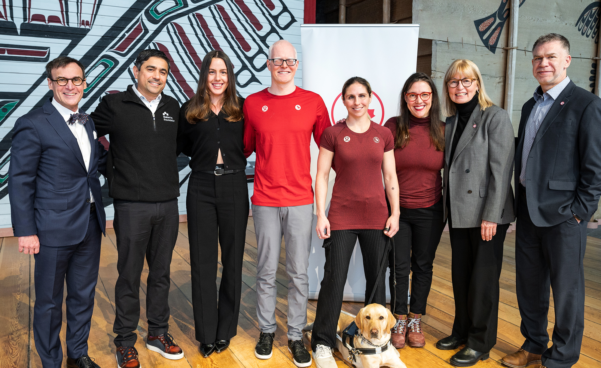 Un groupe de huit personnes, comprenant des athlètes paralympiques, des représentants de fondations et un chien-guide, est debout côte à côte, souriant à la caméra. Le cadre semble être intérieur avec un fond d'œuvres d'art autochtones. Le groupe comprend des hommes et des femmes habillés à la fois en tenue formelle et sportive, mettant en avant un partenariat ou un événement communautaire. Un labrador jaune est assis calmement aux pieds de l'une des personnes.