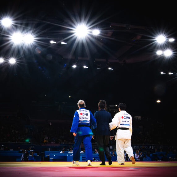 Un judoka malvoyant portant un gi bleu, accompagné par un entraîneur et un officiel, marche vers le centre d'une arène de compétition fortement éclairée sous un projecteur, avec le public en arrière-plan.