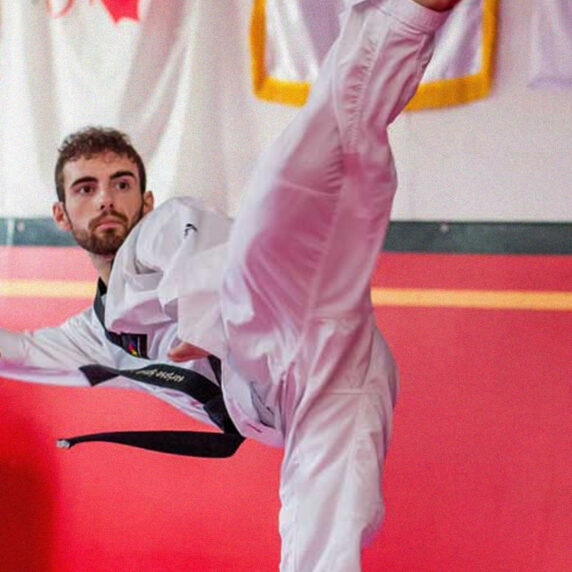 Anthony Cappello, athlète canadien de para-taekwondo, exécute un coup de pied haut dans un dojo, vêtu d’un uniforme blanc de taekwondo et d’une ceinture noire. Les drapeaux du Canada et du Québec sont visibles en arrière-plan, représentant ses origines canadiennes.