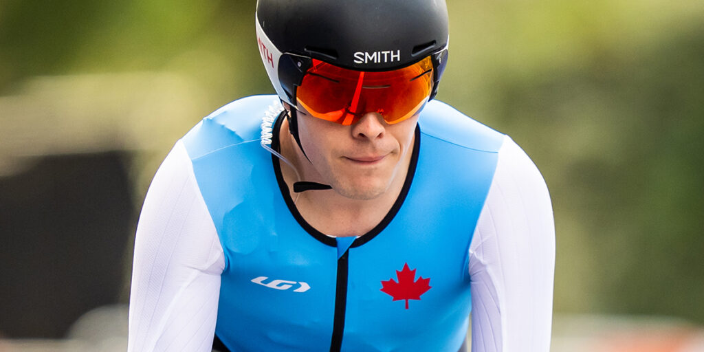 Le Paralympien canadien Nathan Clement roule à toute vitesse sur son vélo, vêtu d’une combinaison de cyclisme bleu clair et blanche ornée d’une feuille d’érable rouge. Son regard concentré et son casque aérodynamique mettent en avant sa détermination pendant la compétition.