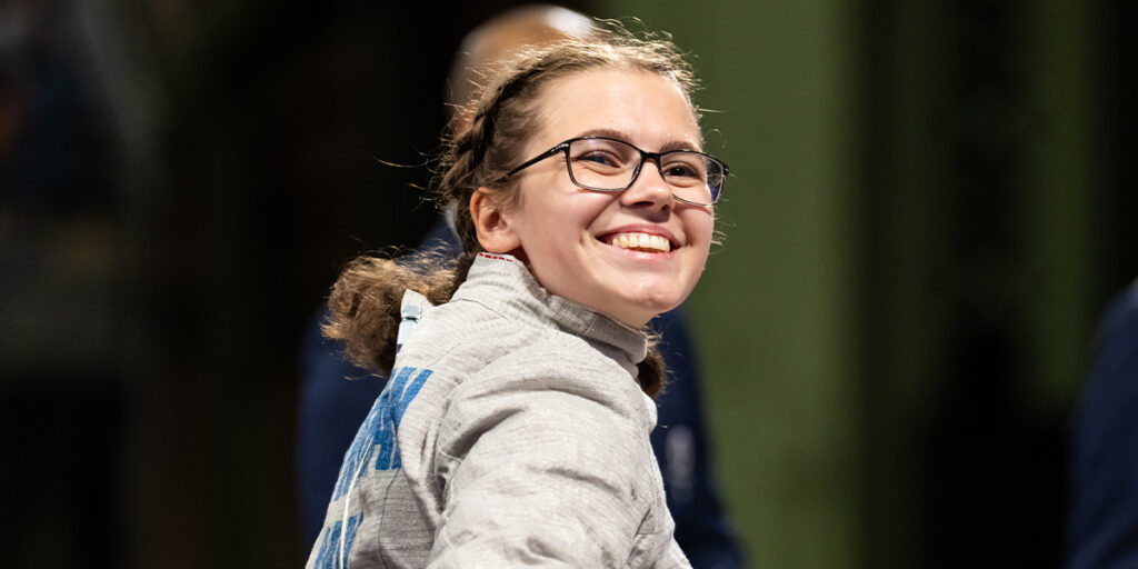 La Paralympienne canadienne Trinity Lowthian sourit avec assurance après un match d’escrime en fauteuil roulant. Elle porte sa veste d’escrime argentée et ses lunettes, tandis que ses cheveux tressés sont soigneusement attachés en arrière. L’atmosphère lumineuse met en valeur ce moment de célébration.