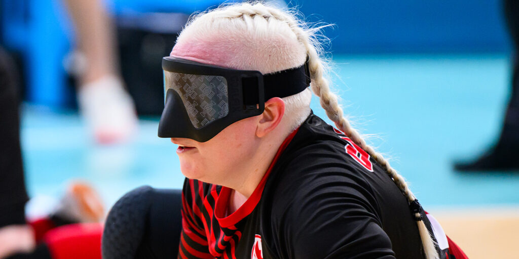 La Paralympienne canadienne Whitney Bogart participe à un match de goalball, vêtue d’un maillot noir et rouge et portant un bandeau sur les yeux, concentrée sur le jeu. Ses cheveux tressés apportent une touche personnelle à son expression déterminée.