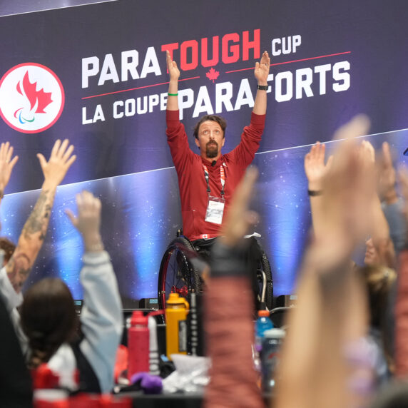 Le paralympien canadien Josh Dueck anime une séance d’échauffement avant un événement de la Coupe ParaForts. Assis dans son fauteuil roulant, Josh lève les bras en hauteur, motivant les participants devant une bannière affichant « ParaTough Cup / La Coupe ParaForts. »