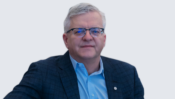 Photo portrait de Richard Rudderham, un homme professionnel aux cheveux gris courts, portant des lunettes, une chemise bleue à col et un blazer foncé. Il arbore une expression calme et confiante, assis devant un fond clair uni.