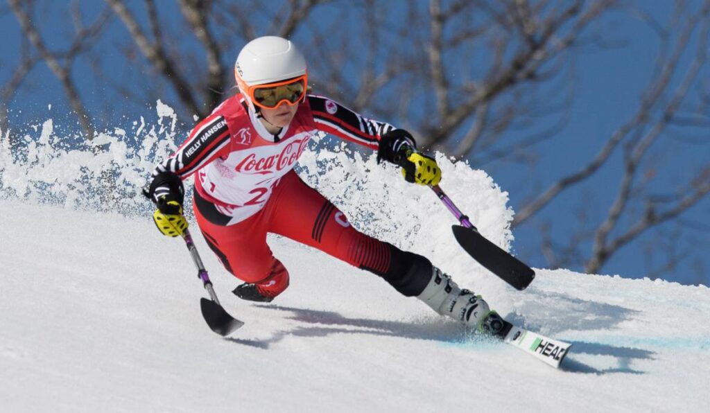 La paralympienne canadienne Frédérique Turgeon participe à une compétition de ski alpin sur une piste enneigée, vêtue d'une combinaison de course rouge et blanche, d'un casque blanc et de gants jaunes. Elle utilise des stabilisateurs pour l'équilibre et la vitesse en taillant dans la neige.