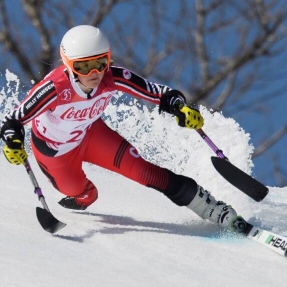 La paralympienne canadienne Frédérique Turgeon participe à une compétition de ski alpin sur une piste enneigée, vêtue d'une combinaison de course rouge et blanche, d'un casque blanc et de gants jaunes. Elle utilise des stabilisateurs pour l'équilibre et la vitesse en taillant dans la neige.