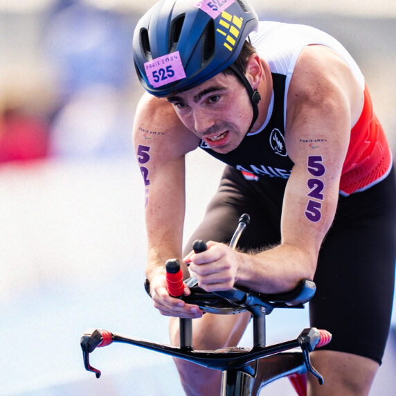 Un athlète déterminé portant une combinaison de triathlon canadienne participe à une course à vélo spécialisée lors des Jeux paralympiques de Paris 2024. Le numéro 525 est visible sur ses bras et son casque, symbolisant son esprit compétitif.