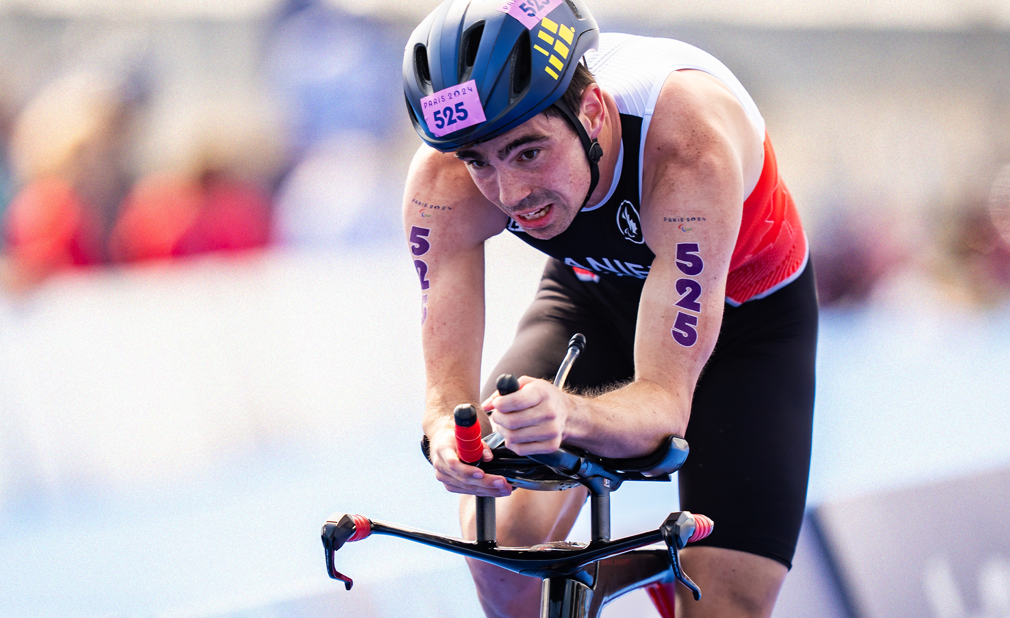 Un athlète déterminé portant une combinaison de triathlon canadienne participe à une course à vélo spécialisée lors des Jeux paralympiques de Paris 2024. Le numéro 525 est visible sur ses bras et son casque, symbolisant son esprit compétitif.