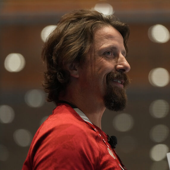 Vue de profil du médaillé d’or paralympique canadien Josh Dueck souriant, portant un chandail rouge et s’exprimant sur scène avec un micro-casque.