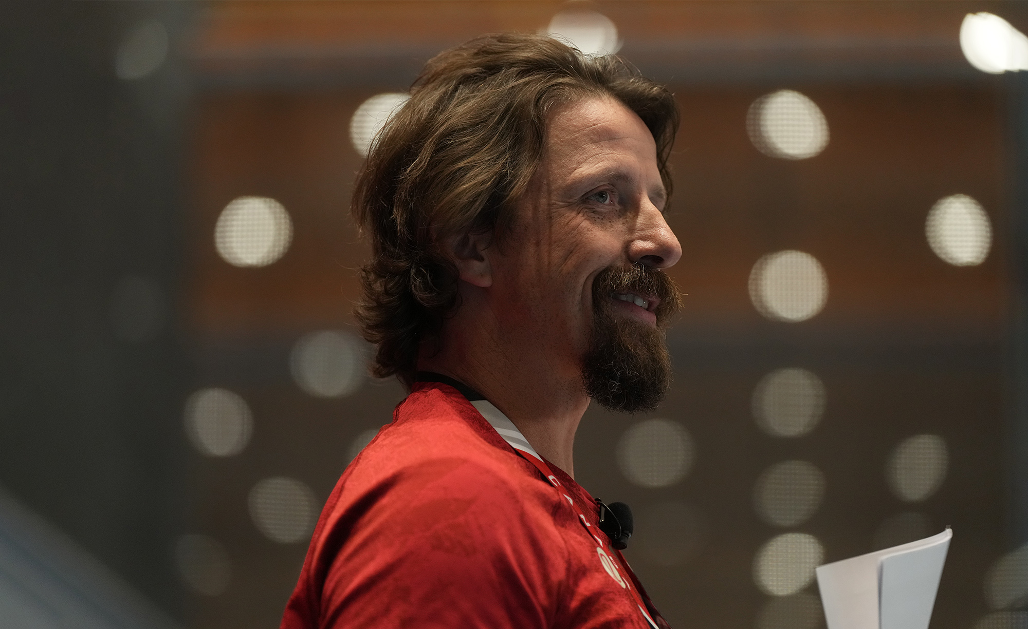 Vue de profil du médaillé d’or paralympique canadien Josh Dueck souriant, portant un chandail rouge et s’exprimant sur scène avec un micro-casque.