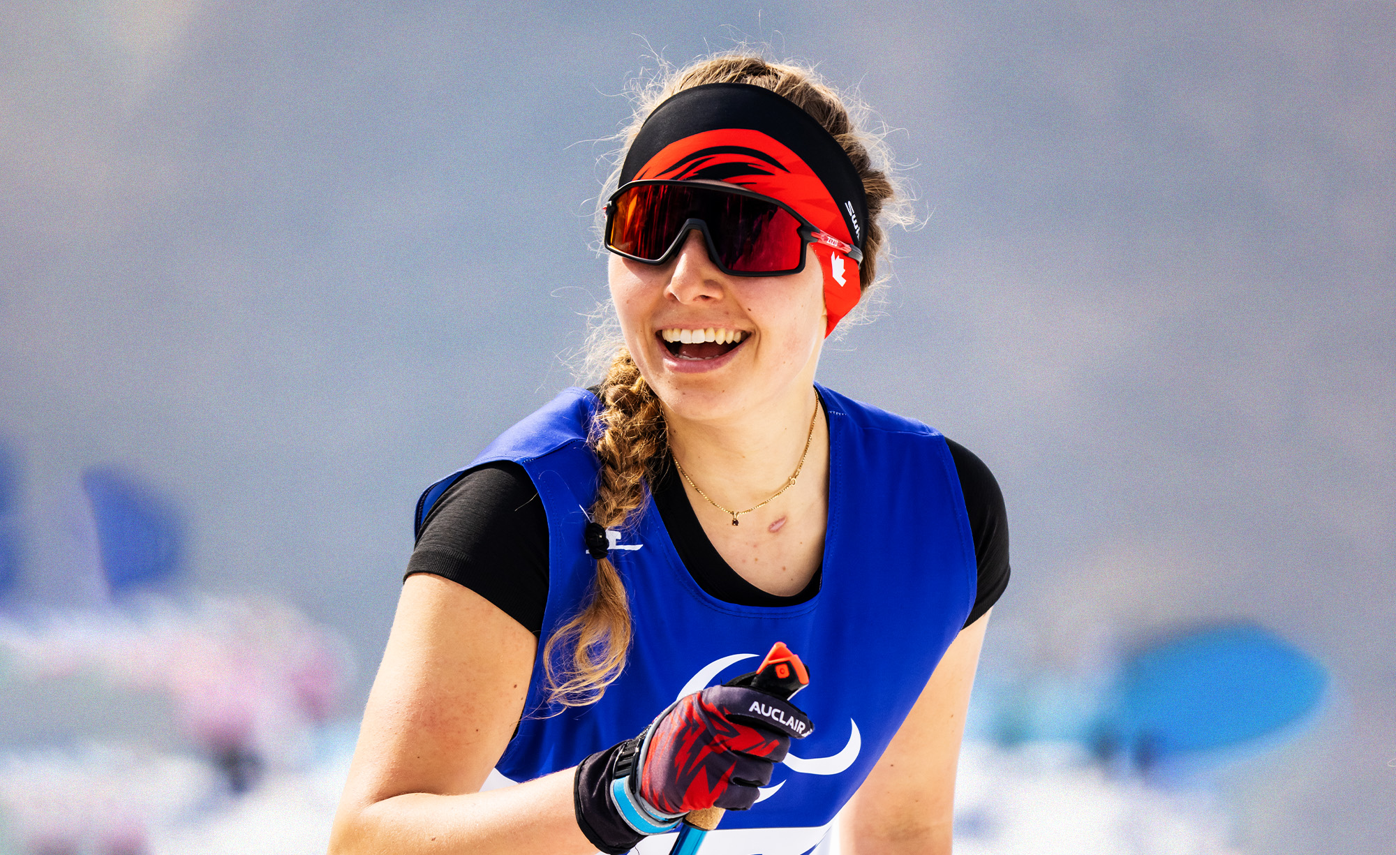 La paralympienne canadienne Natalie Wilkie sourit largement pendant une compétition de ski paranordique. Elle porte une chasuble de course bleue, un bandeau rouge et des lunettes de soleil, tenant son bâton de ski en main. L’arrière-plan enneigé est flou.