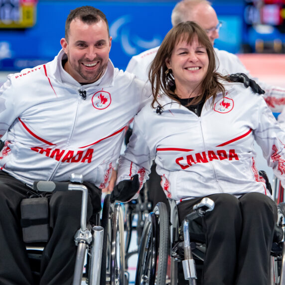 Les curleurs canadiens en fauteuil roulant Jon Thurston et Ina Forrest sourient ensemble sur la glace, portant des vestes blanches avec des accents de feuille d'érable rouge et le mot « CANADA » imprimé sur leur poitrine. Leurs coéquipiers sont visibles en arrière-plan pendant un match de curling.