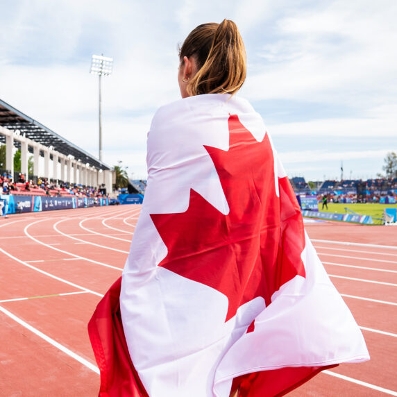 Keegan Gaunt, Paralympienne canadienne, enveloppée dans le drapeau du Canada, debout sur la piste lors des Jeux parapanaméricains de Santiago 2023. Le stade est rempli de spectateurs sous un ciel dégagé.