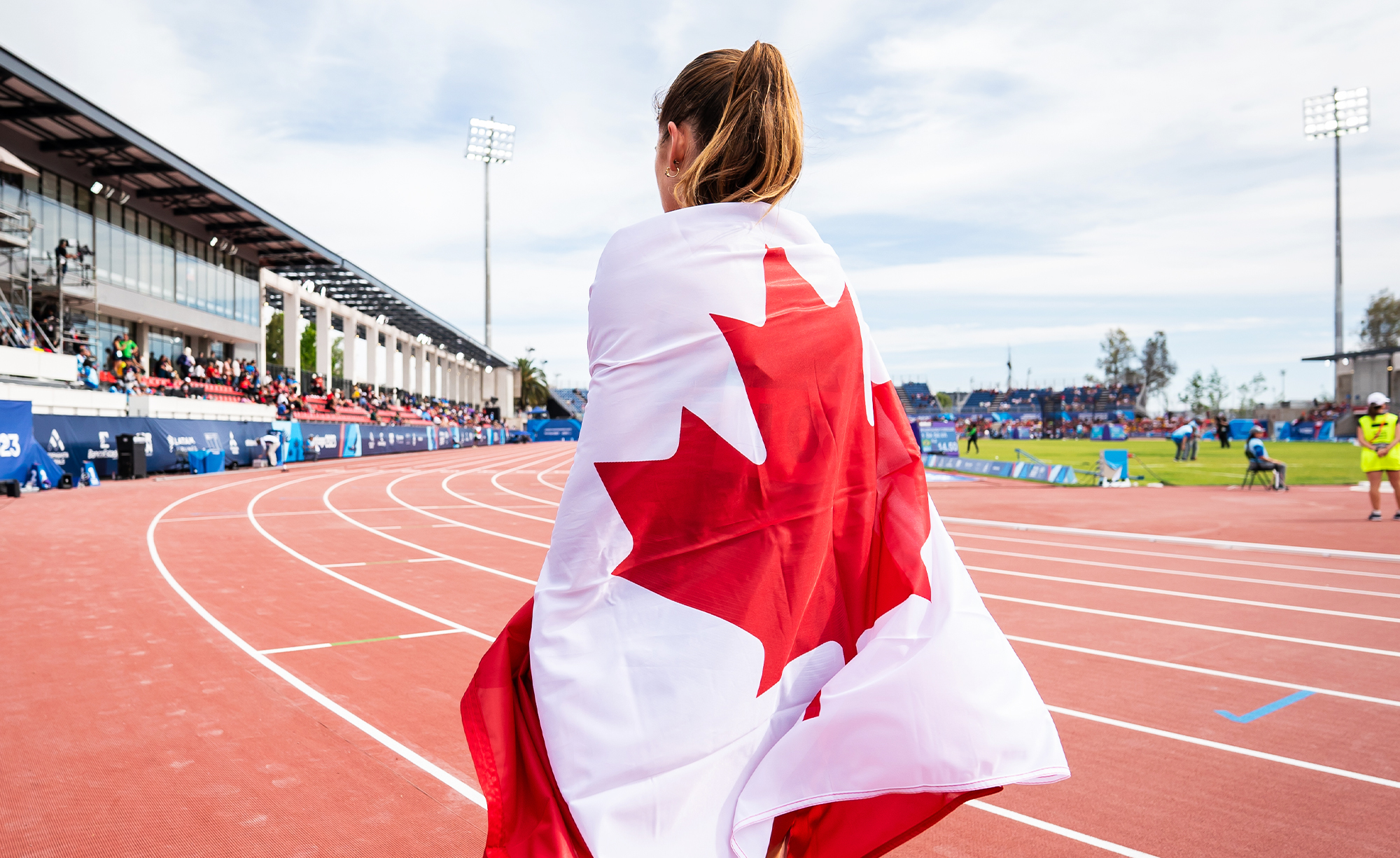 Keegan Gaunt, Paralympienne canadienne, enveloppée dans le drapeau du Canada, debout sur la piste lors des Jeux parapanaméricains de Santiago 2023. Le stade est rempli de spectateurs sous un ciel dégagé.