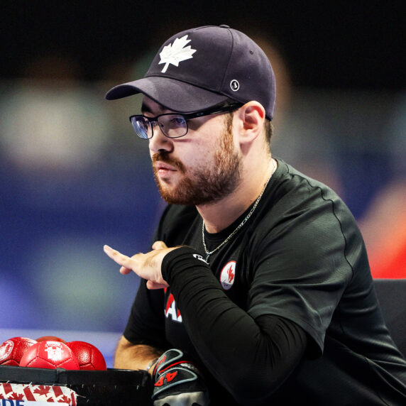 Le paralympien canadien de boccia Danik Allard est concentré pendant un match. Il porte un chandail noir et une casquette noire ornée d’une feuille d’érable blanche, et est assis à côté de balles de boccia rouges dans une boîte aux couleurs du Canada. L’arrière-plan est flou avec des couleurs vives.