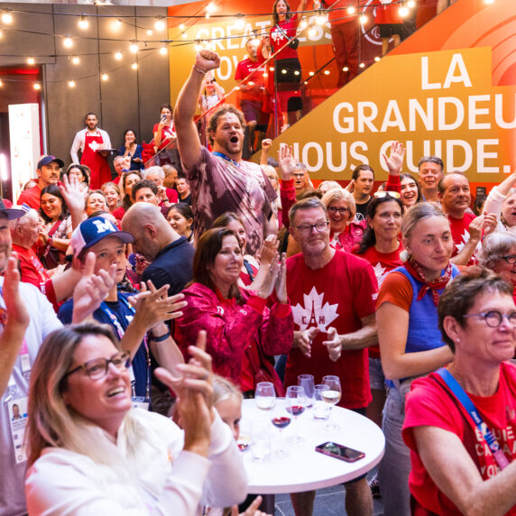 Une foule animée de partisans canadiens vêtus de rouge et blanc applaudit avec enthousiasme lors de l'événement à la Maison Paralympique Canadienne pendant les Jeux paralympiques de Paris 2024. Le lieu est décoré de branding canadien, incluant une bannière indiquant 'La Grandeur Nous Guide.' L'ambiance est festive, avec des guirlandes lumineuses suspendues et des sourires partout.