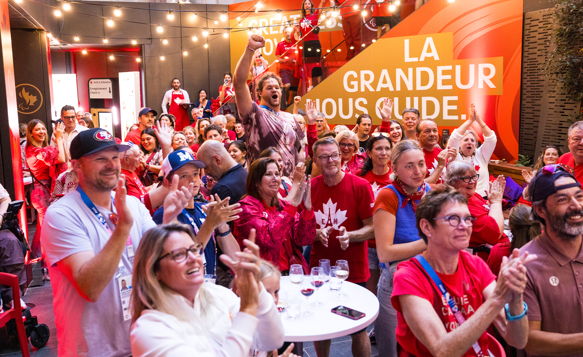 Une foule animée de partisans canadiens vêtus de rouge et blanc applaudit avec enthousiasme lors de l'événement à la Maison Paralympique Canadienne pendant les Jeux paralympiques de Paris 2024. Le lieu est décoré de branding canadien, incluant une bannière indiquant 'La Grandeur Nous Guide.' L'ambiance est festive, avec des guirlandes lumineuses suspendues et des sourires partout.