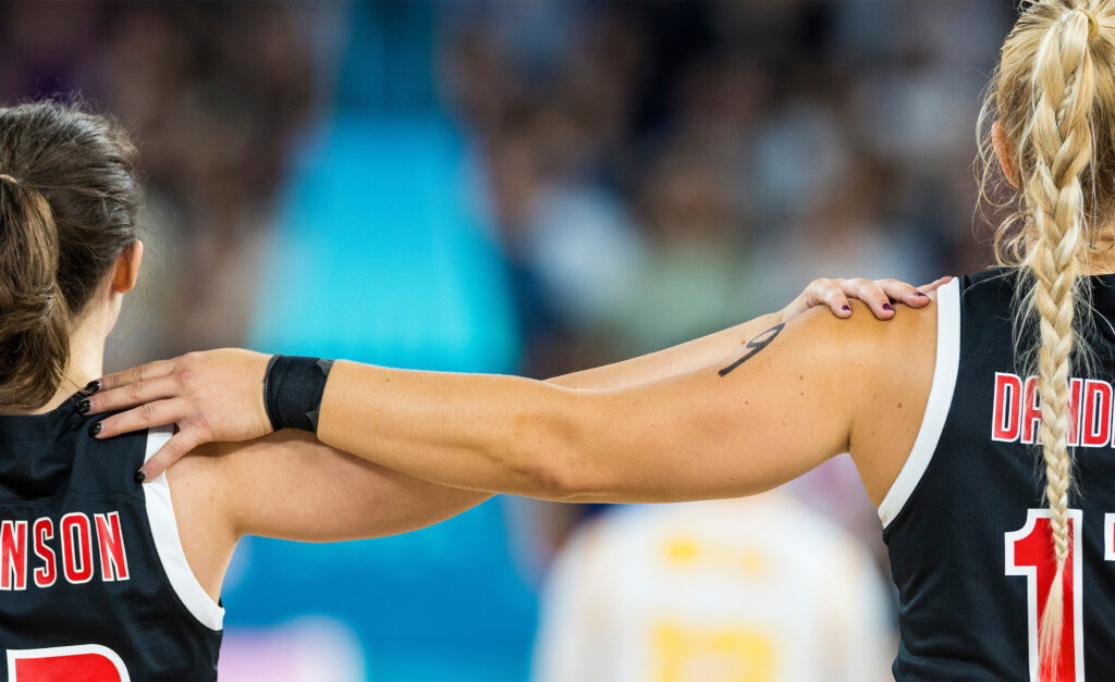 Plan rapproché de deux athlètes canadiennes en maillots noirs avec des lettres rouges et blanches, debout épaule contre épaule avec les bras autour l'une de l'autre. L'athlète à droite a une coiffure tressée et le numéro 11 sur son maillot.