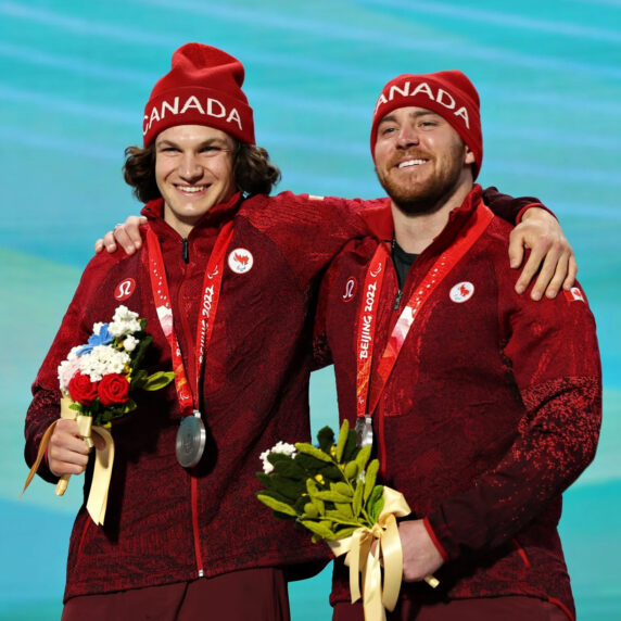 Mac Marcoux (à droite) se tient sur le podium aux Jeux paralympiques de Beijing 2022, vêtu d’une veste rouge de l’équipe du Canada et d’un bonnet assorti, tenant un bouquet de fleurs. Il sourit aux côtés de son guide, Tristan Rodgers (à gauche), alors qu’ils célèbrent leur médaille d’argent dans l’épreuve de descente hommes, catégorie malvoyants.