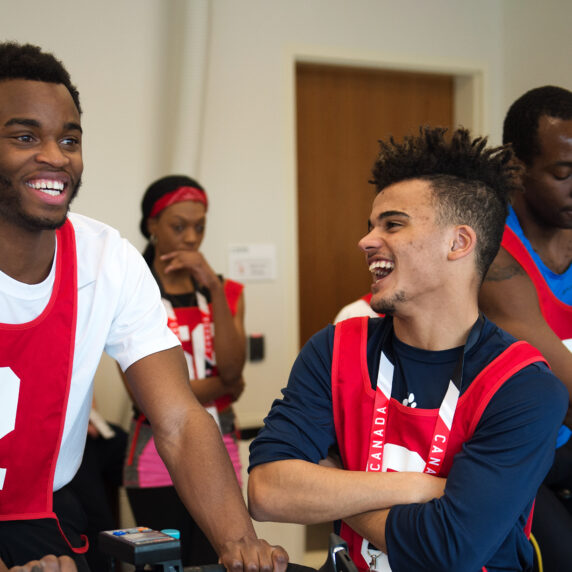 Deux jeunes hommes portant des chasubles rouges numérotées rient ensemble pendant une activité de cyclisme intérieur. En arrière-plan, d'autres participants vêtus de tenues similaires participent à des activités, créant une ambiance vivante et énergique.