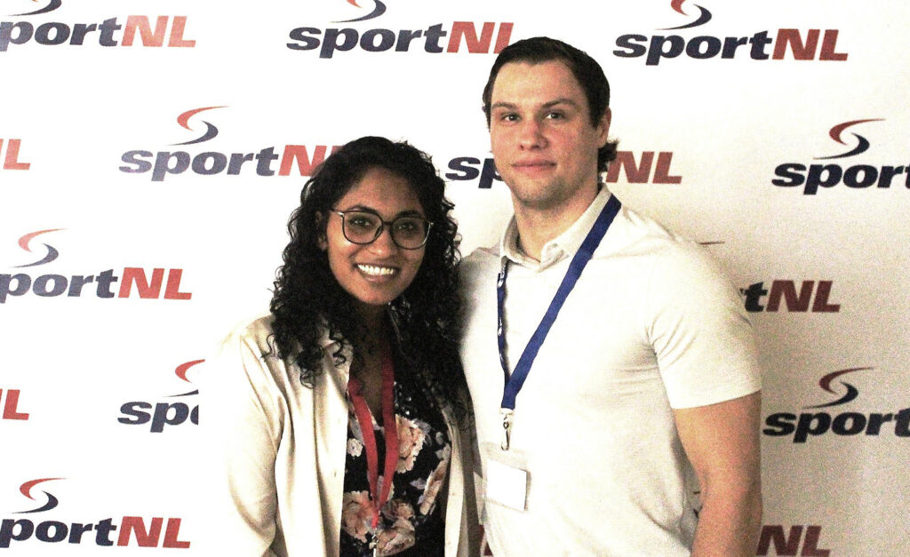 Deux personnes souriantes posent devant un arrière-plan SportNL. La femme à gauche porte des lunettes, un haut à motif floral et un blazer clair, tandis que l'homme à droite porte une chemise blanche à col et un cordon bleu.