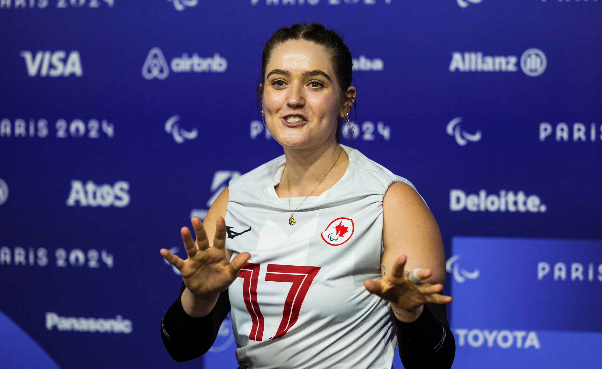 La joueuse canadienne de volley-ball assis Allison Lang, portant un maillot blanc d’Équipe Canada avec le numéro 17, sourit lors d’une entrevue d’après-match avec un journaliste de CBC aux Jeux paralympiques de Paris 2024. L’arrière-plan présente un mur médiatique bleu avec le logo des Jeux paralympiques de Paris 2024 et des logos de partenaires.