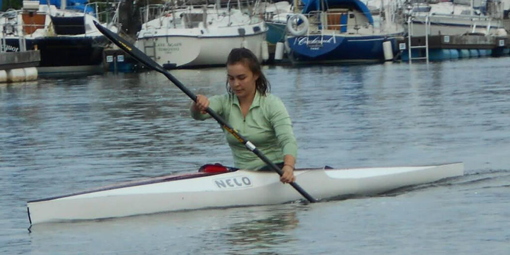 La paralympienne canadienne Erica Scarff pagaie dans un kayak blanc de marque Nelo sur des eaux calmes, portant une chemise verte à manches longues. Plusieurs voiliers sont amarrés en arrière-plan dans une marina.