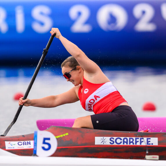 La paralympienne canadienne Erica Scarff participe à une épreuve de para canoë aux Jeux paralympiques de Paris 2024. Elle porte un uniforme rouge et blanc de l’équipe canadienne et pagaie avec puissance dans son embarcation rouge et noire, marquée de son nom de famille, « Scarff E. ». Le numéro de course « 5 » est visible sur son bateau. Une grande bannière « Paris 2024 » apparaît en arrière-plan flou.