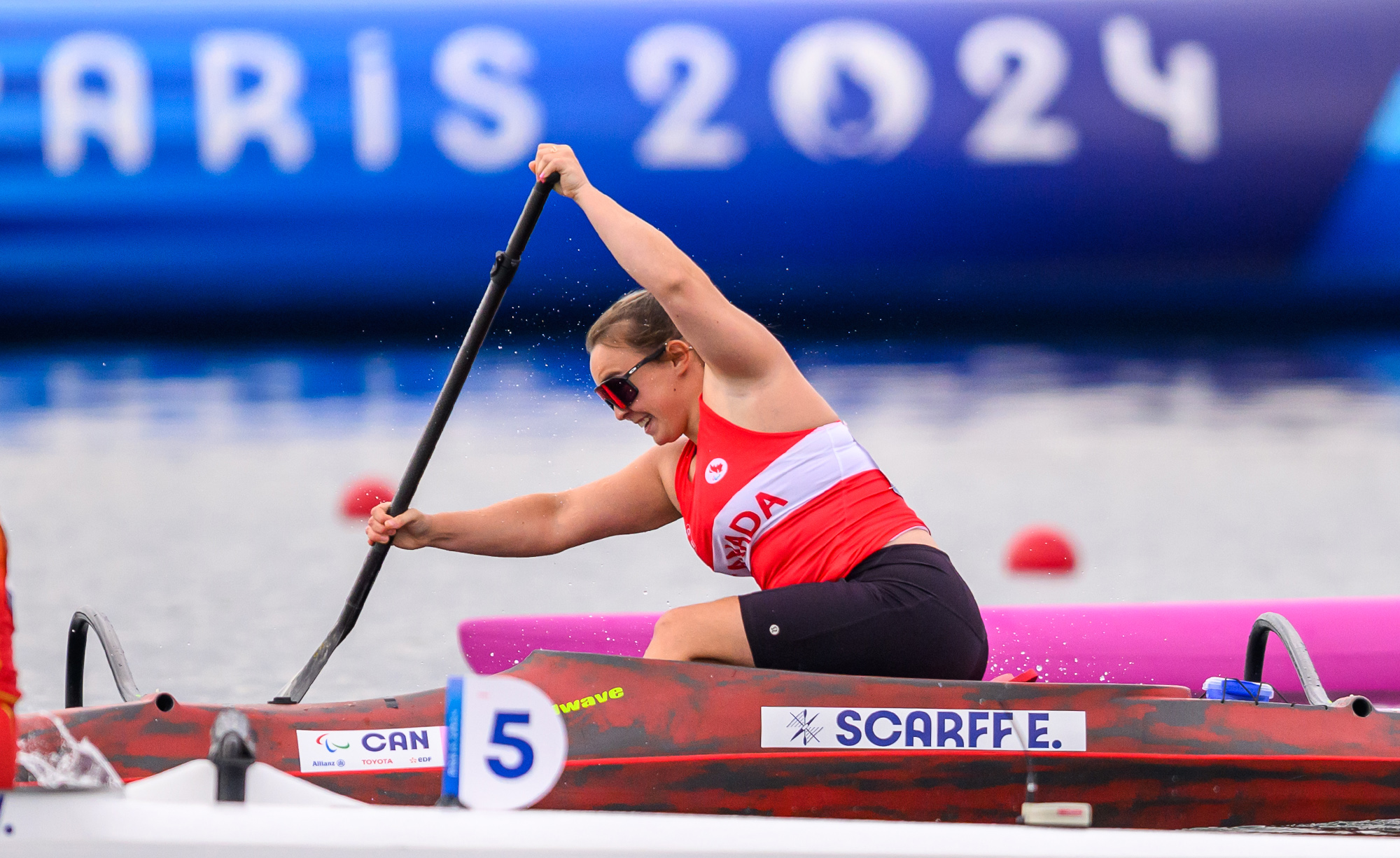 La paralympienne canadienne Erica Scarff participe à une épreuve de para canoë aux Jeux paralympiques de Paris 2024. Elle porte un uniforme rouge et blanc de l’équipe canadienne et pagaie avec puissance dans son embarcation rouge et noire, marquée de son nom de famille, « Scarff E. ». Le numéro de course « 5 » est visible sur son bateau. Une grande bannière « Paris 2024 » apparaît en arrière-plan flou.