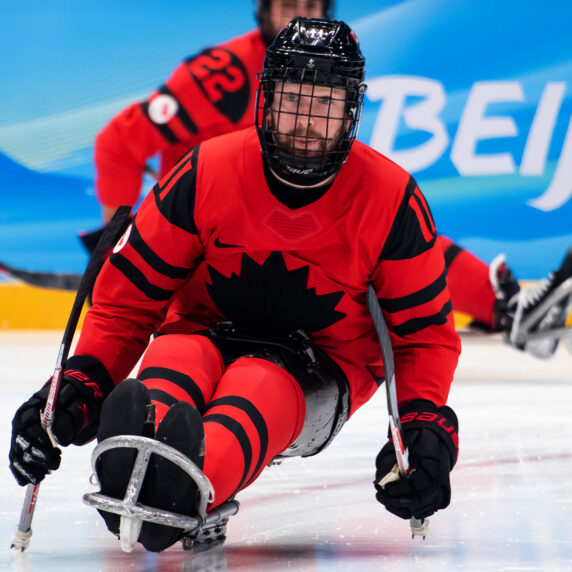 Un joueur de hockey sur luge de l’équipe paralympique canadienne, portant un chandail rouge et noir avec un emblème de feuille d’érable noire, participe aux Jeux paralympiques d’hiver de Beijing 2022. Il est assis dans une luge, tenant deux bâtons de hockey munis de pics en métal pour la propulsion. Un autre joueur canadien est visible en arrière-plan sur la glace, avec le mot « Beijing » affiché sur les panneaux bleus de l’aréna.