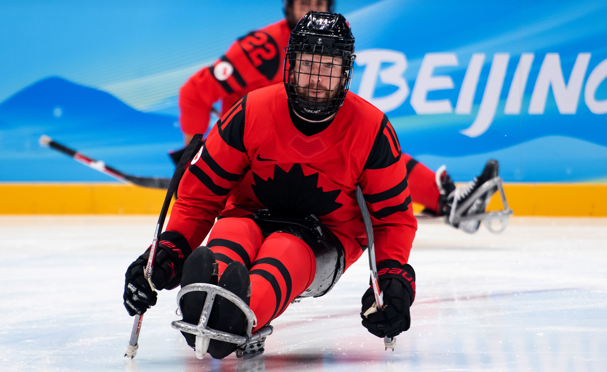 Un joueur de hockey sur luge de l’équipe paralympique canadienne, portant un chandail rouge et noir avec un emblème de feuille d’érable noire, participe aux Jeux paralympiques d’hiver de Beijing 2022. Il est assis dans une luge, tenant deux bâtons de hockey munis de pics en métal pour la propulsion. Un autre joueur canadien est visible en arrière-plan sur la glace, avec le mot « Beijing » affiché sur les panneaux bleus de l’aréna.