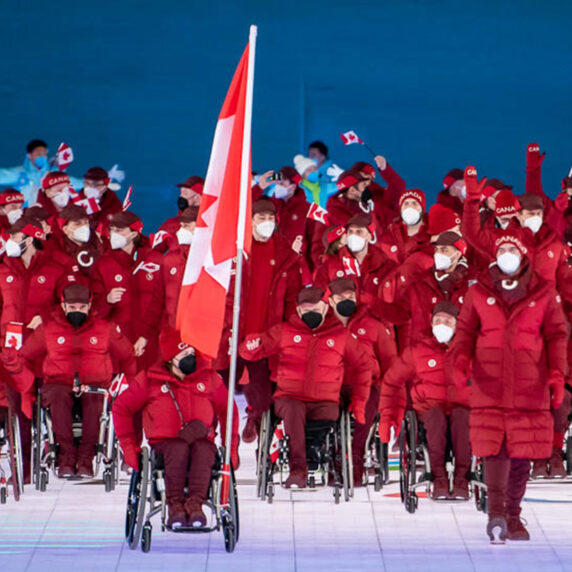 Les athlètes de l’équipe paralympique canadienne, vêtus de vestes et de tuques rouges assorties, entrent fièrement dans le stade lors de la cérémonie d’ouverture des Jeux paralympiques. Certains athlètes agitent des drapeaux canadiens, tandis que d’autres avancent en fauteuil roulant. Un grand drapeau canadien est porté à l’avant du groupe. Tous les athlètes portent des masques faciaux blancs et l’arrière-plan présente un éclairage bleu avec une grande lanterne rouge.