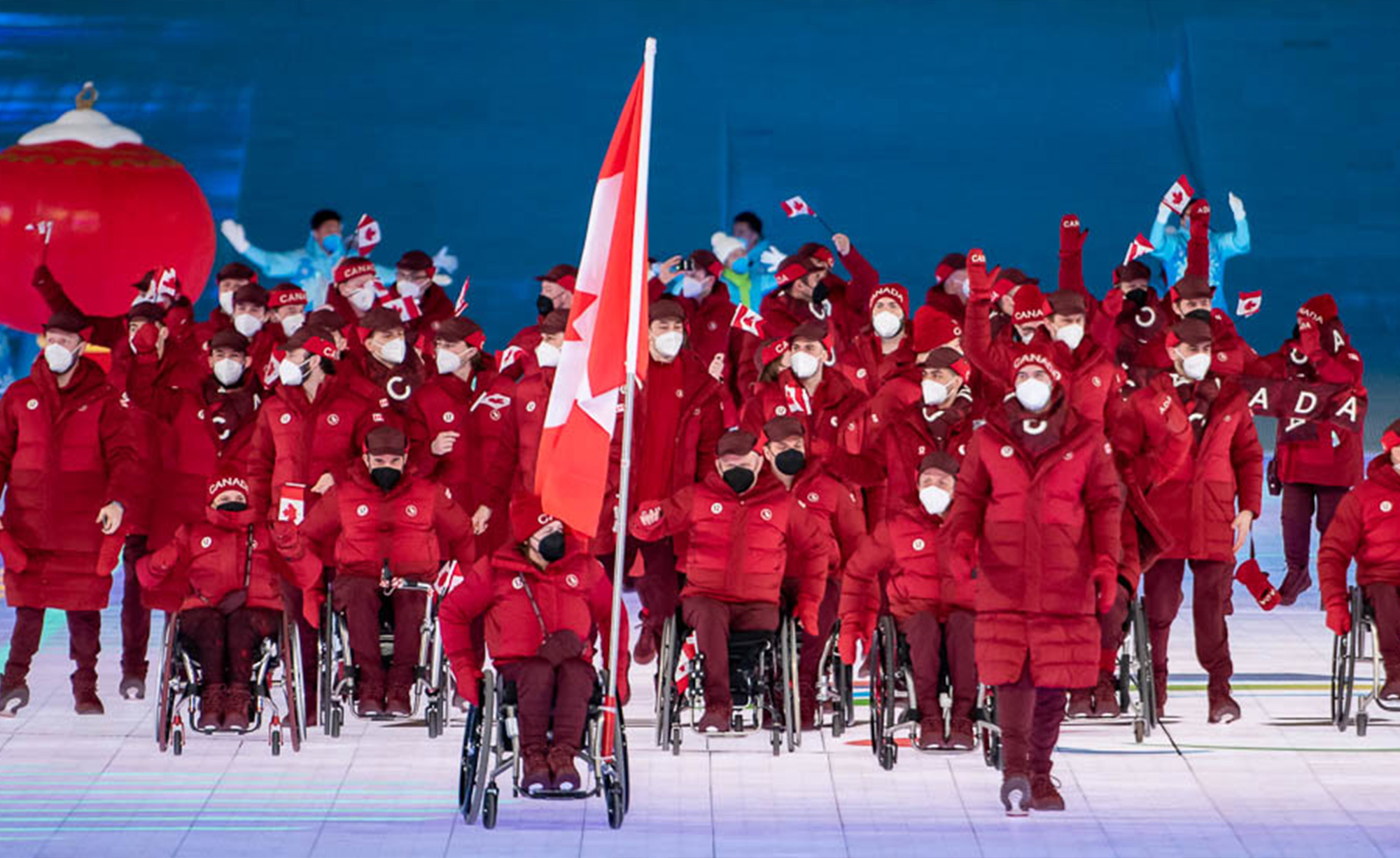 Les athlètes de l’équipe paralympique canadienne, vêtus de vestes et de tuques rouges assorties, entrent fièrement dans le stade lors de la cérémonie d’ouverture des Jeux paralympiques. Certains athlètes agitent des drapeaux canadiens, tandis que d’autres avancent en fauteuil roulant. Un grand drapeau canadien est porté à l’avant du groupe. Tous les athlètes portent des masques faciaux blancs et l’arrière-plan présente un éclairage bleu avec une grande lanterne rouge.